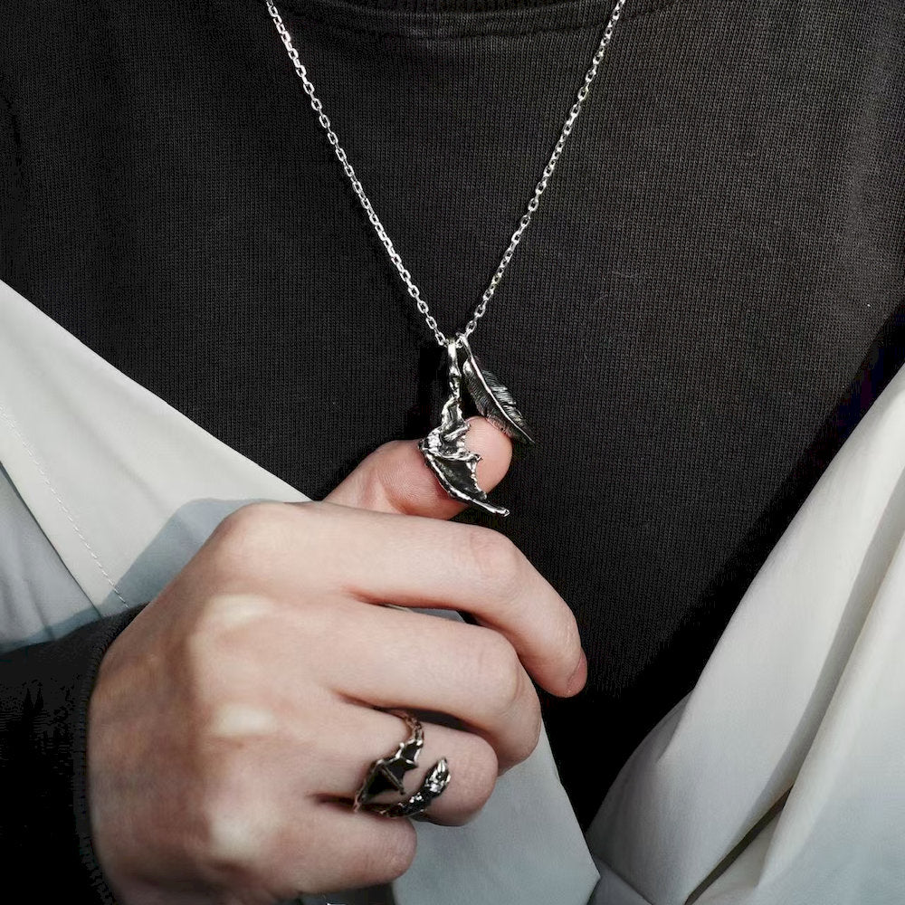 Hand holding an oxidized silver necklace with a dragon wing pendant against a black background